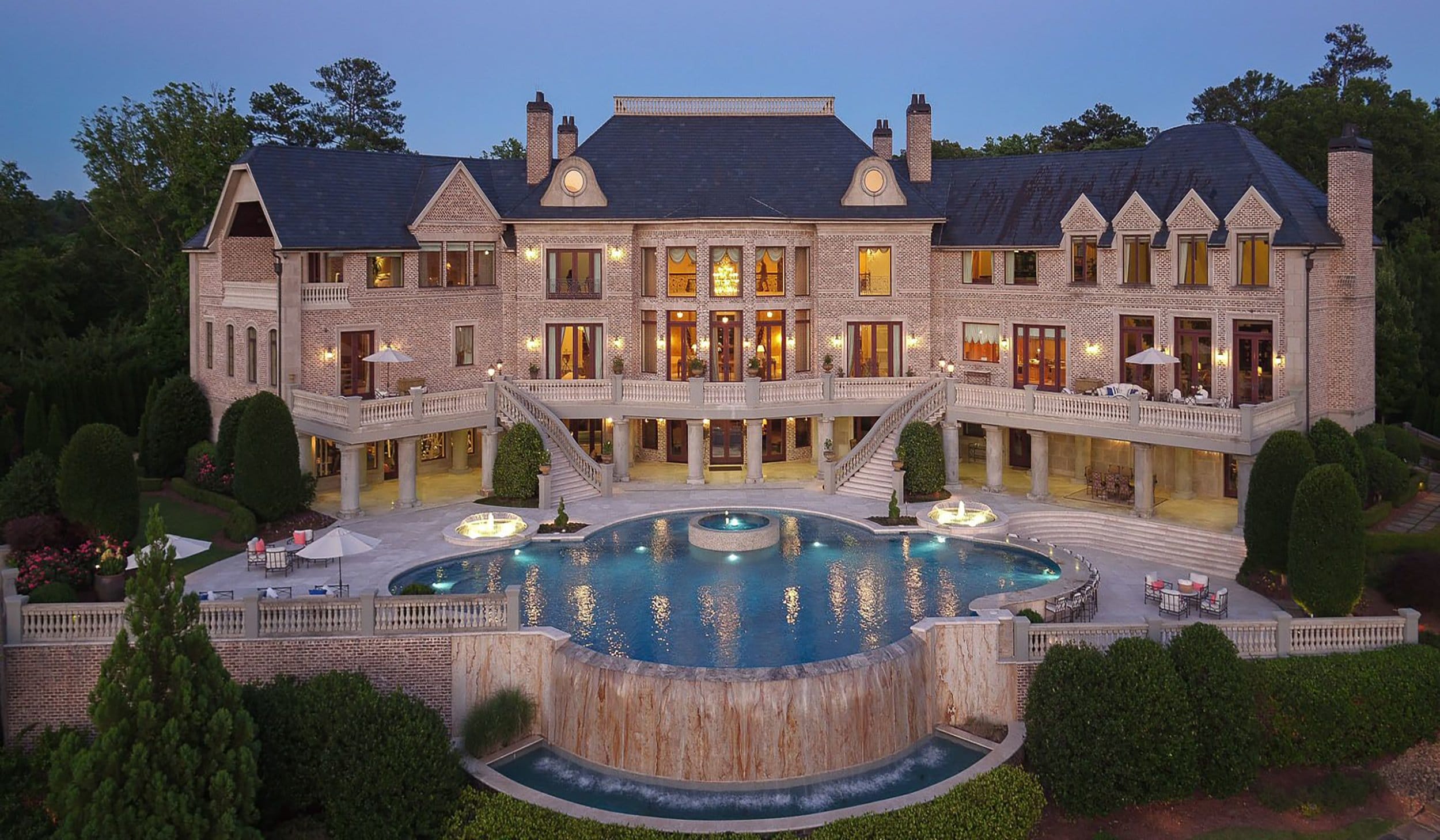 Luxurious mansion with grand staircase, large windows, and a stunning pool in the foreground surrounded by lush greenery.