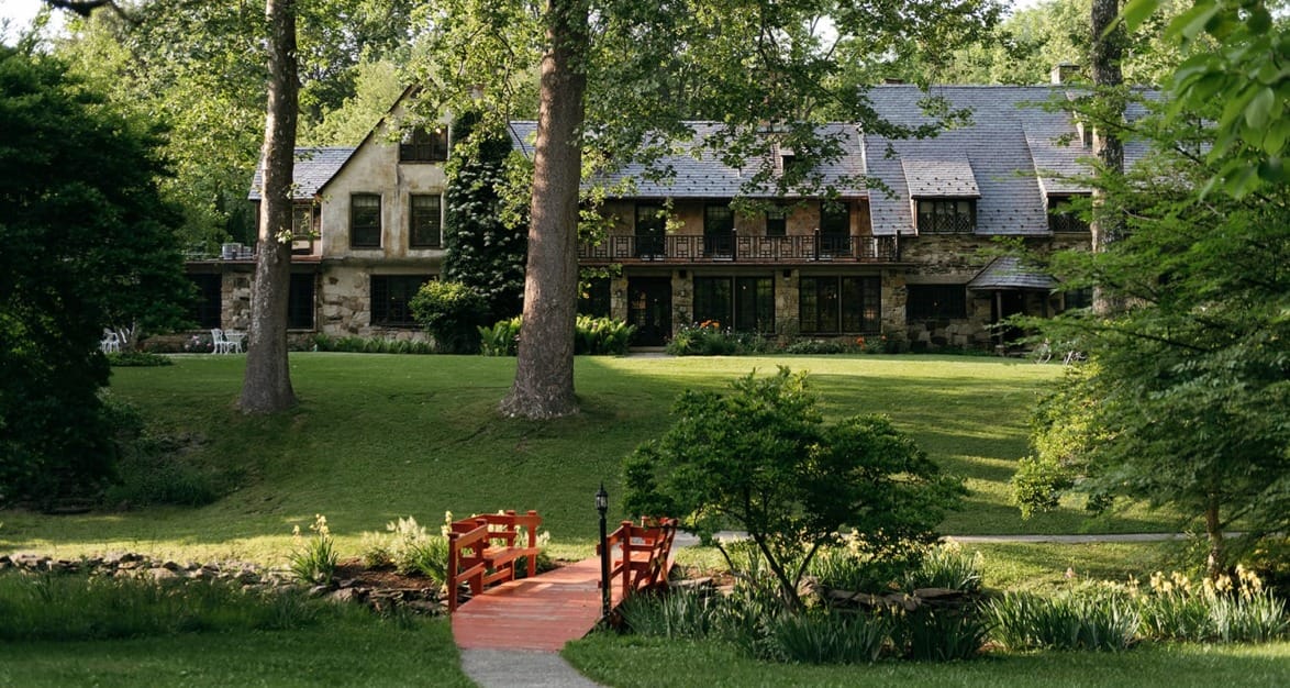 A large rustic house with a slate roof and lush green lawn, featuring a small red wooden bridge in the foreground.