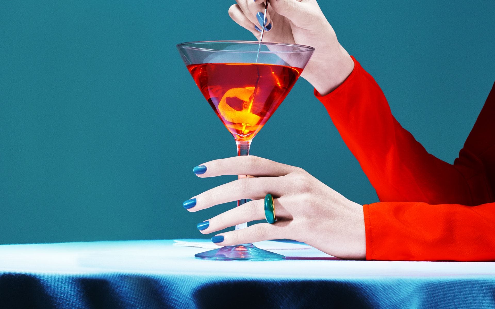 Person stirring a vibrant red cocktail with a lemon slice, wearing a red sleeve and blue nail polish, set against a teal background.