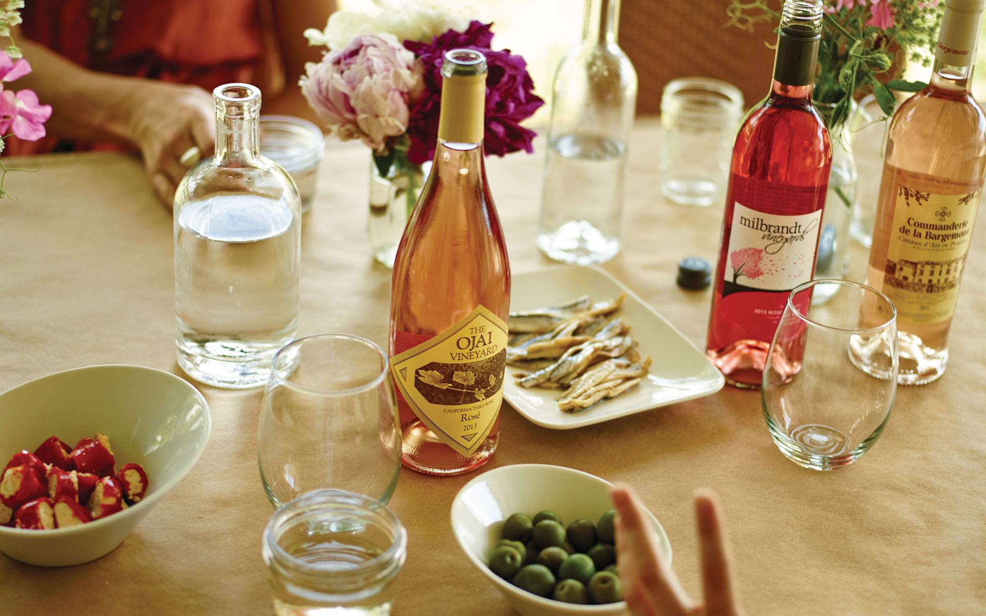 Table with various wine bottles, glassware, water, olives, and snacks, adorned with flower arrangements.