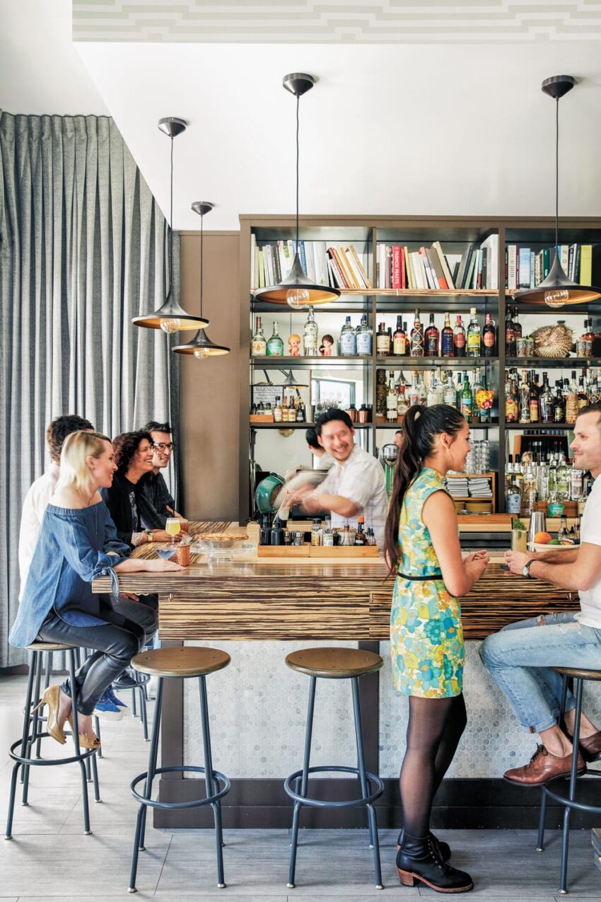 People enjoying drinks at a lively bar with a bartender mixing cocktails and a shelf filled with bottles and books in the background.