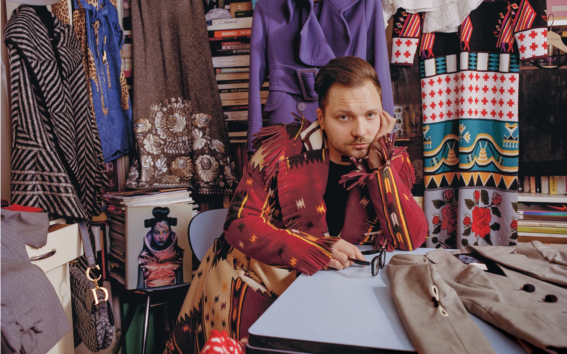 Fashion designer sitting at a desk surrounded by colorful, patterned clothing and books.