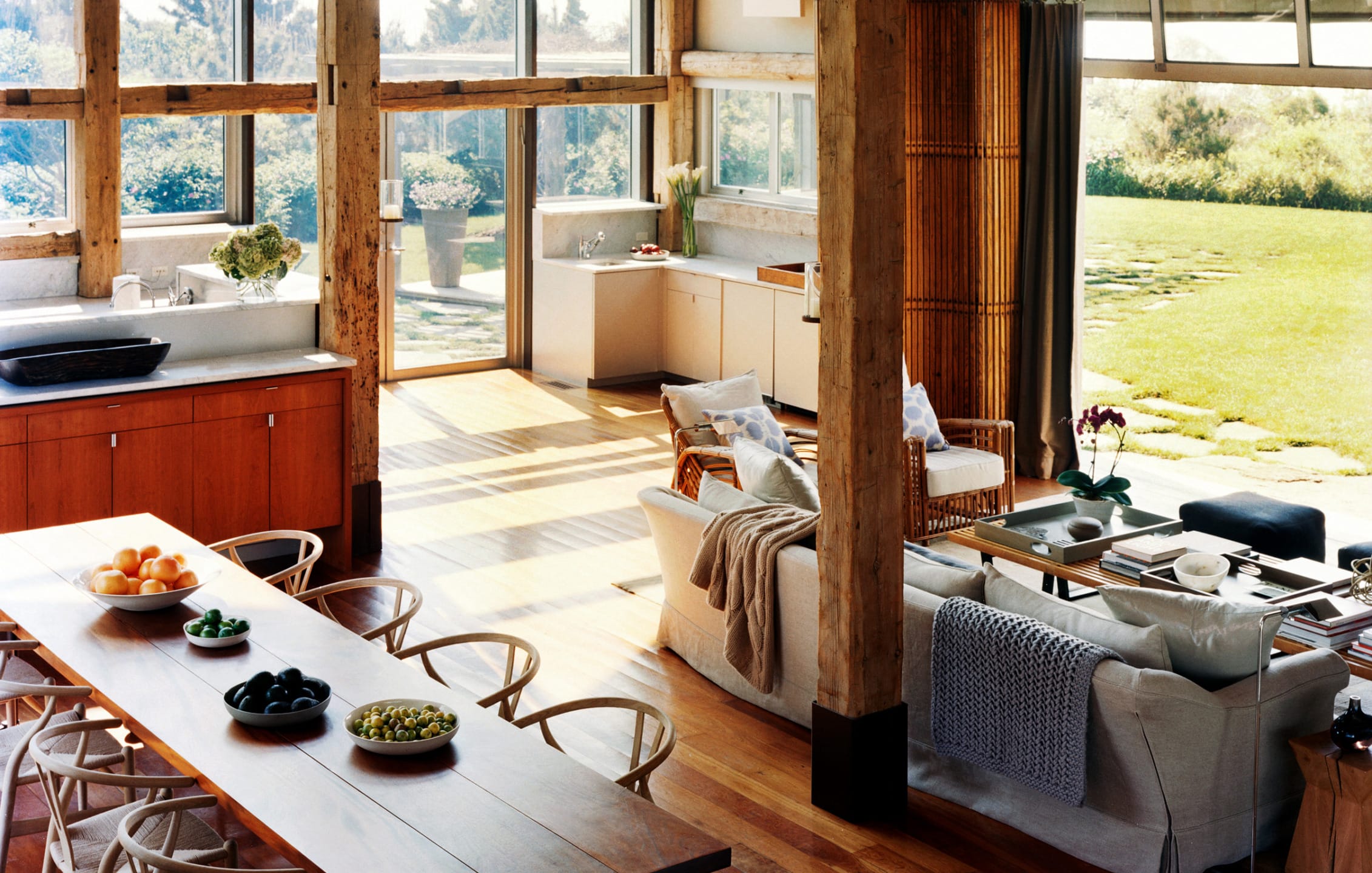 Open-concept living room and kitchen with wooden beams, large windows, dining table, sofas, and garden view.