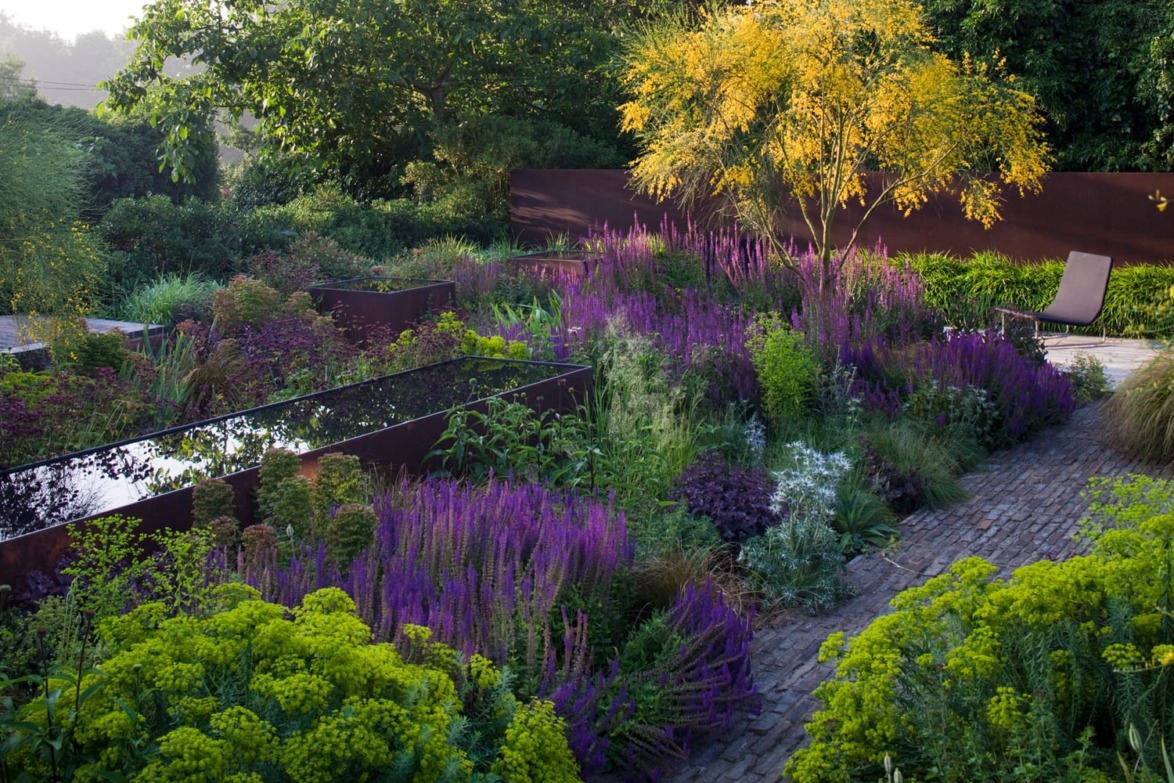 A lush garden with purple and yellow flowers, greenery, and a reflective water feature, surrounded by trees and a brick path.