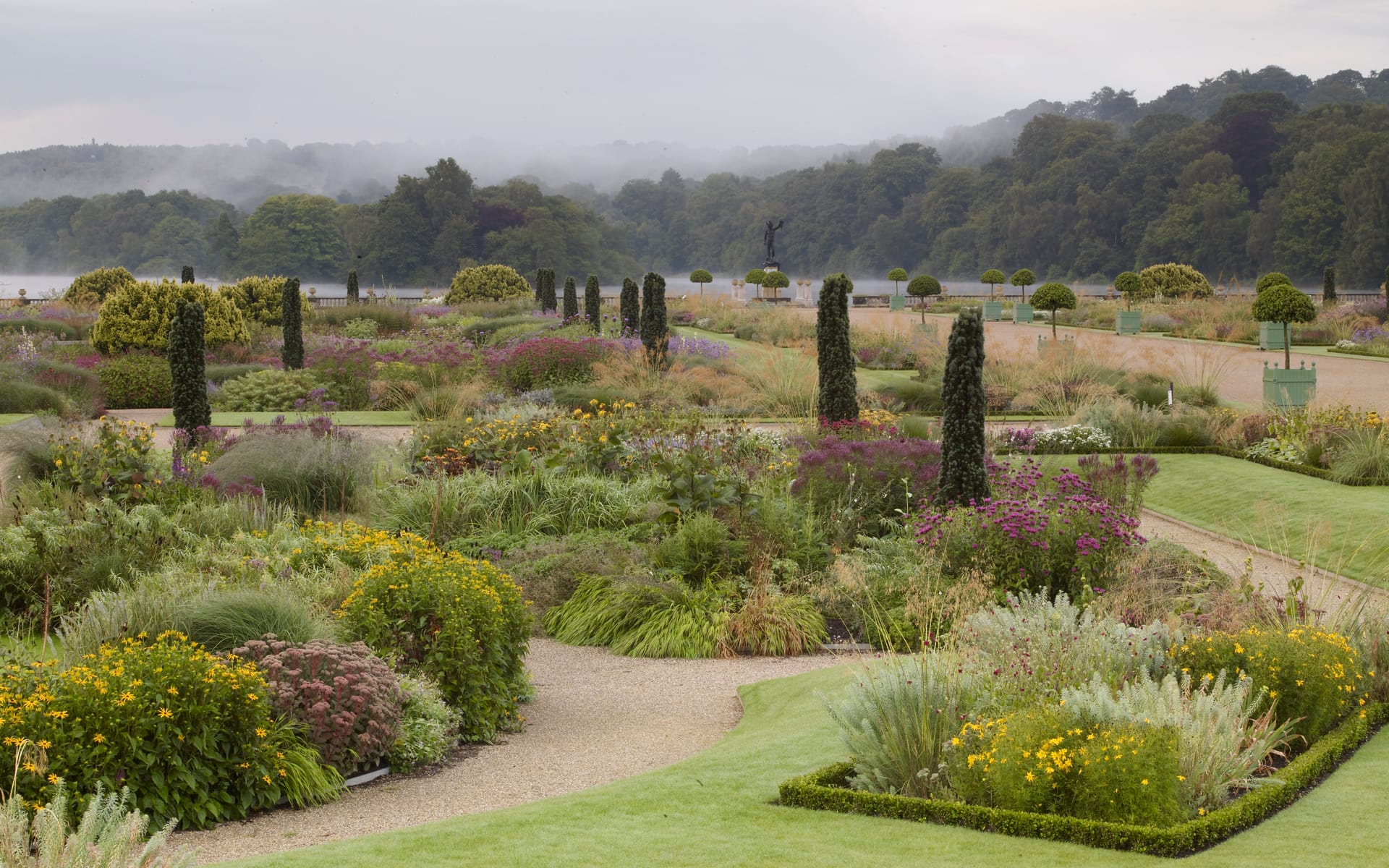 Vibrant formal garden with diverse flowers, manicured hedges, and misty trees in the background on a cloudy day.
