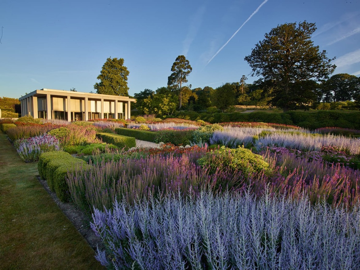 Botanic garden with purple and green shrubs, neatly trimmed hedges, and a modern building in the background under a clear sky.