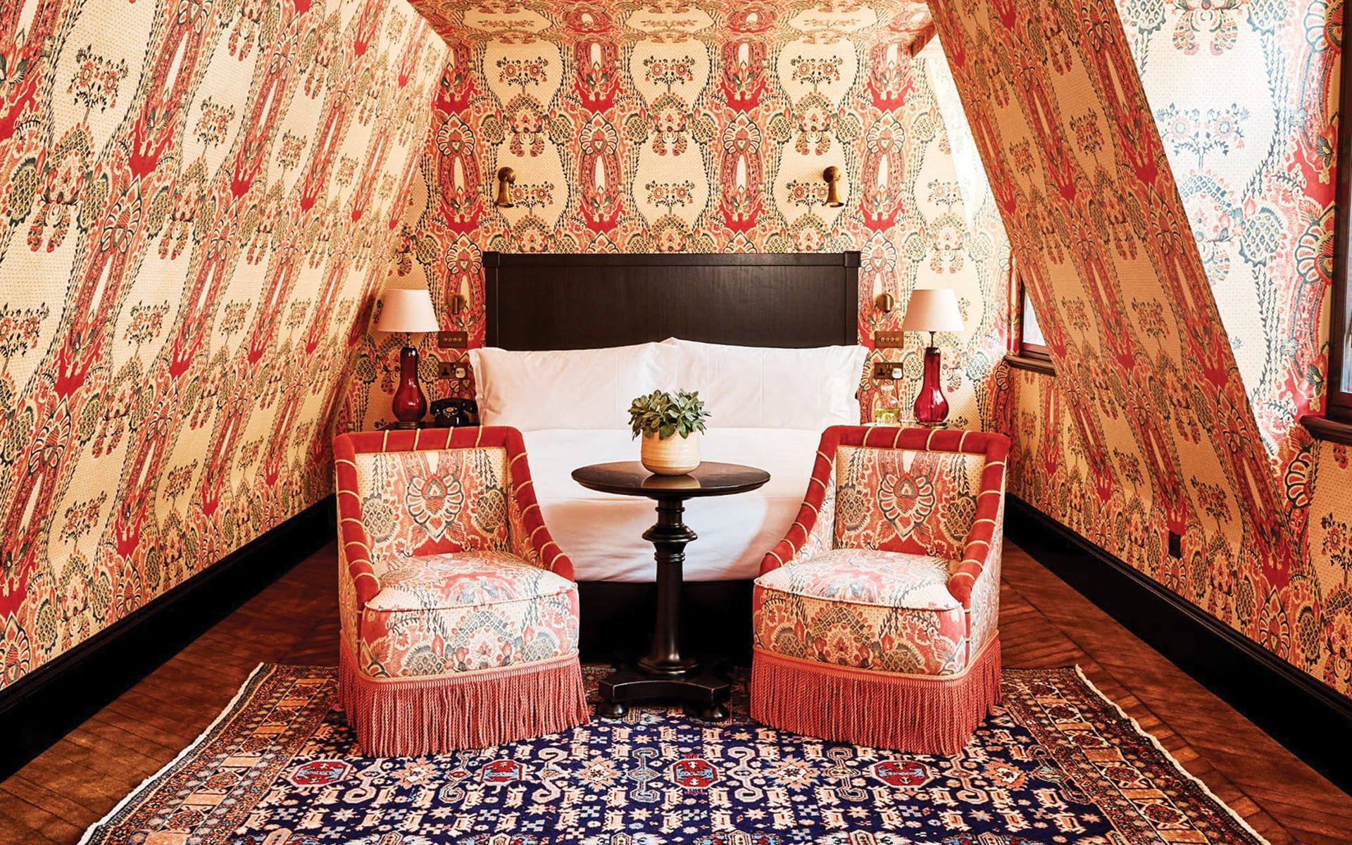 Cozy attic bedroom with patterned wallpaper, two red chairs, a small table, and a bed on a decorative rug.