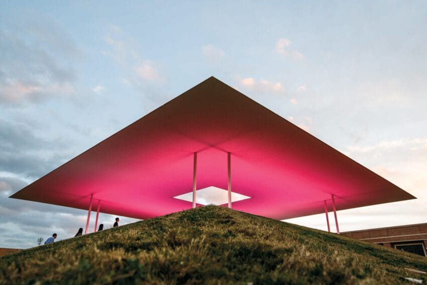 People walking under a large geometric pavilion with a glowing pink light during sunset.