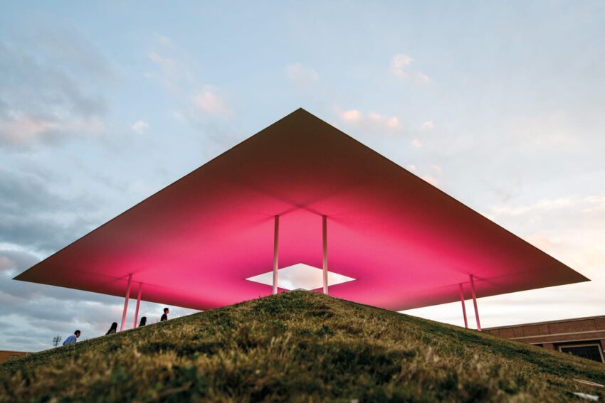 People walking under a large geometric pavilion with a glowing pink light during sunset.