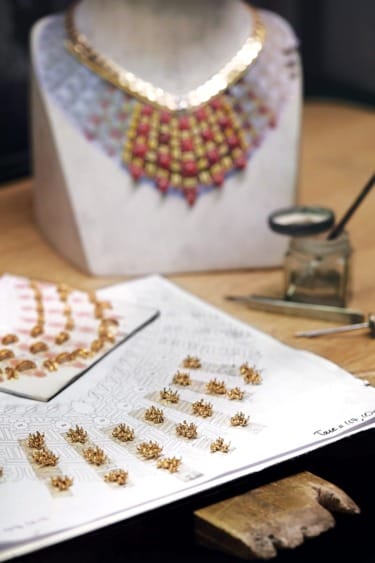 Jewelry design process with gold components on paper, tools, and completed necklace displayed on a stand.