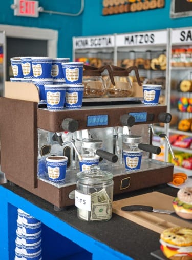 Coffee machine with blue paper cups, tip jar with money in front, bagels in the background.