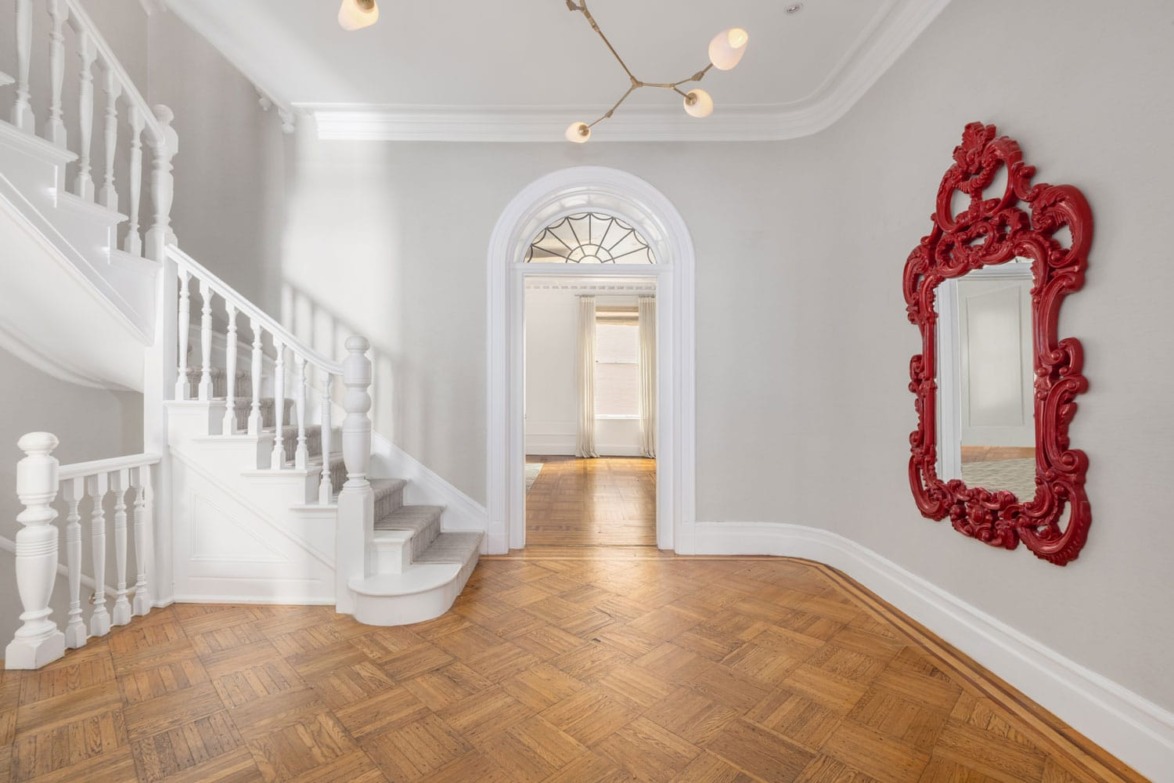 Elegant hallway with parquet floor, white staircase, ornate red mirror, and modern lighting fixture.