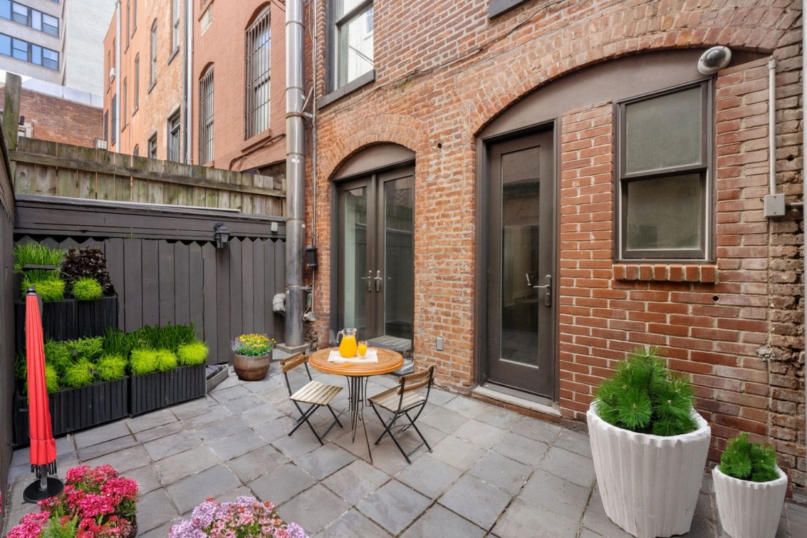 Cozy patio with a small table and chairs, surrounded by potted plants, red umbrella, and brick building backdrop.
