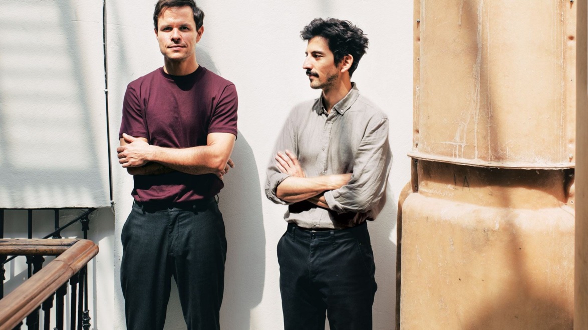 Two men standing on a sunlit balcony, one in a burgundy shirt and the other in a gray shirt, both with arms crossed.