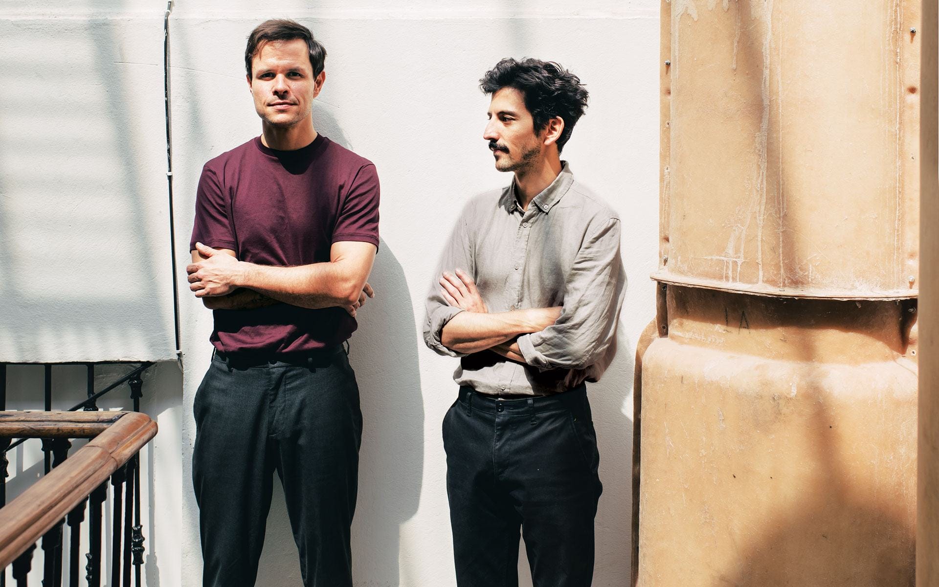 Two men standing on a sunlit balcony, one in a burgundy shirt and the other in a gray shirt, both with arms crossed.