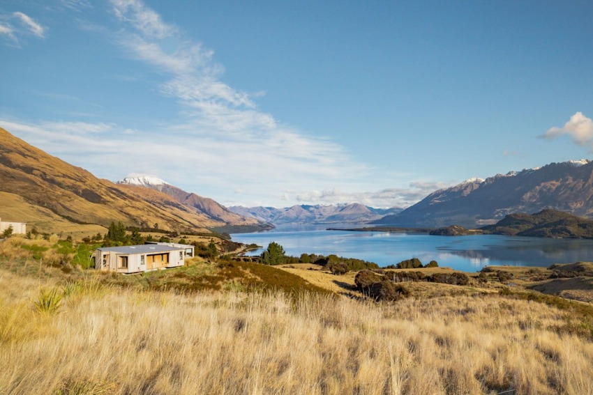 Scenic view of a house in a grassy field by a serene lake surrounded by mountains under a clear blue sky.
