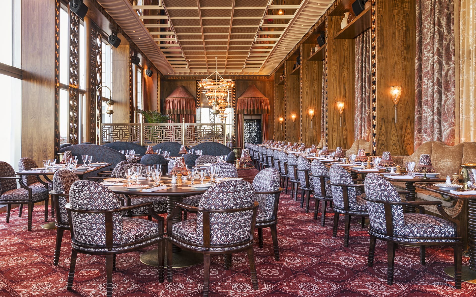Elegant restaurant interior with patterned chairs, set dining tables, ornate wooden panels, and large windows.