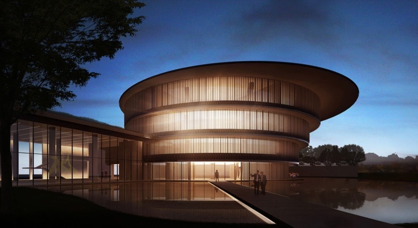 Modern circular building illuminated at night, reflecting on a still pond with trees in the background.