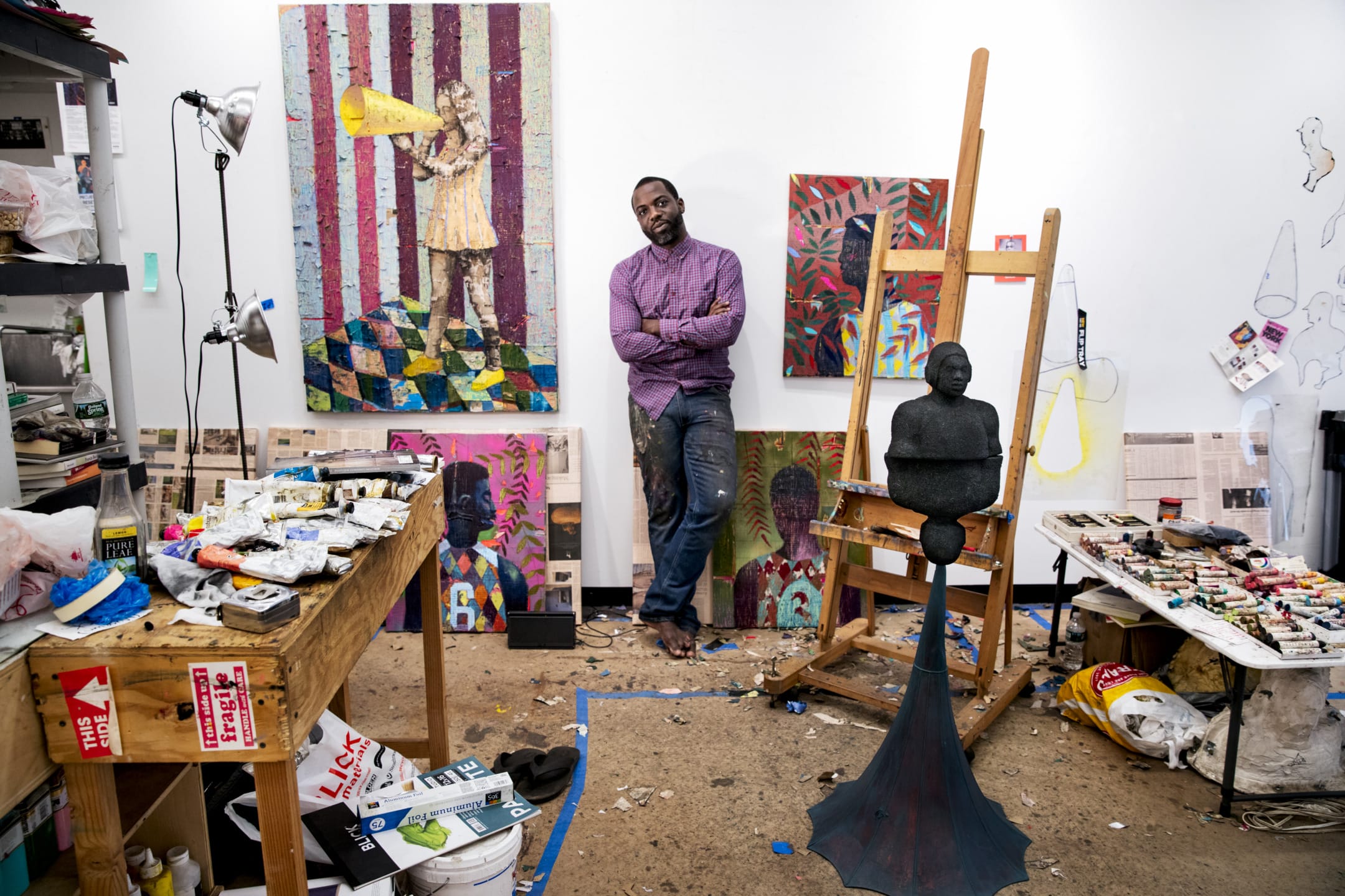 Artist standing in a colorful studio surrounded by paintings, art supplies, and a sculpture on an easel.