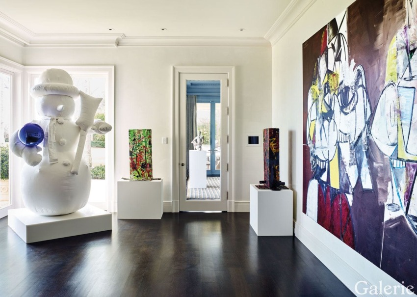 Modern art gallery room with a large white balloon sculpture, abstract paintings, and colorful pedestal displays on dark wood floor.