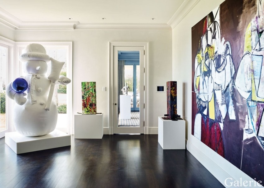 Modern art gallery room with a large white balloon sculpture, abstract paintings, and colorful pedestal displays on dark wood floor.