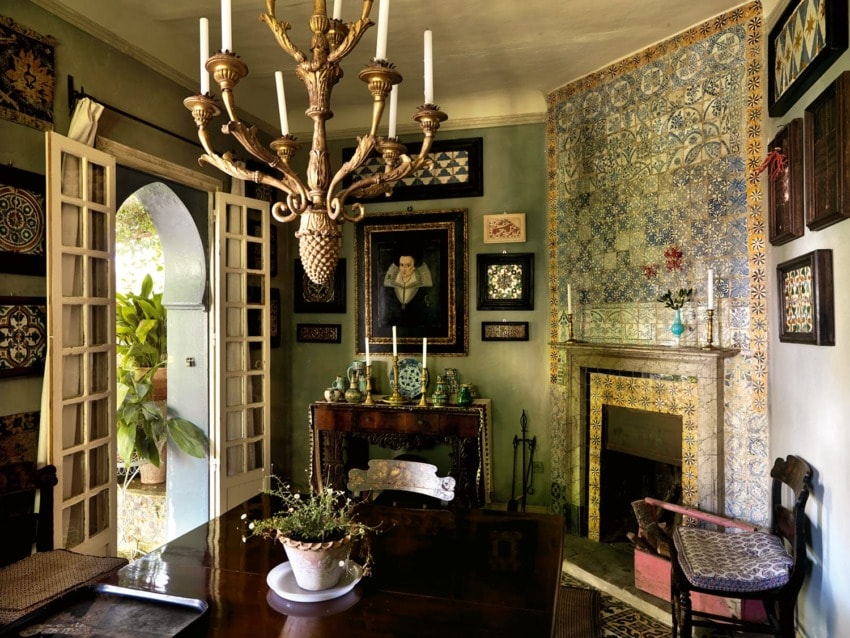 Elegant dining room with ornate chandelier, intricate tiled fireplace, artwork, and greenery through open French doors.