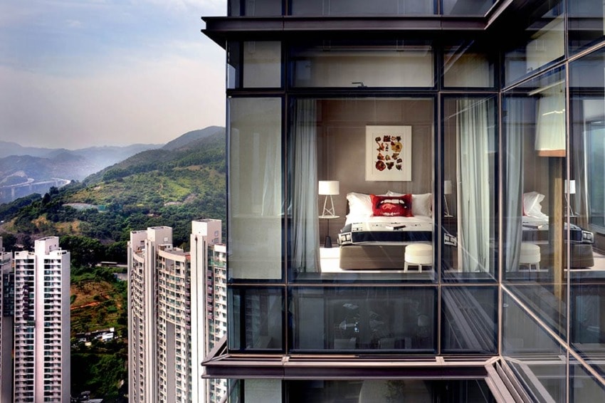 Modern glass-walled bedroom in high-rise, overlooking a cityscape with mountains and greenery in the background.