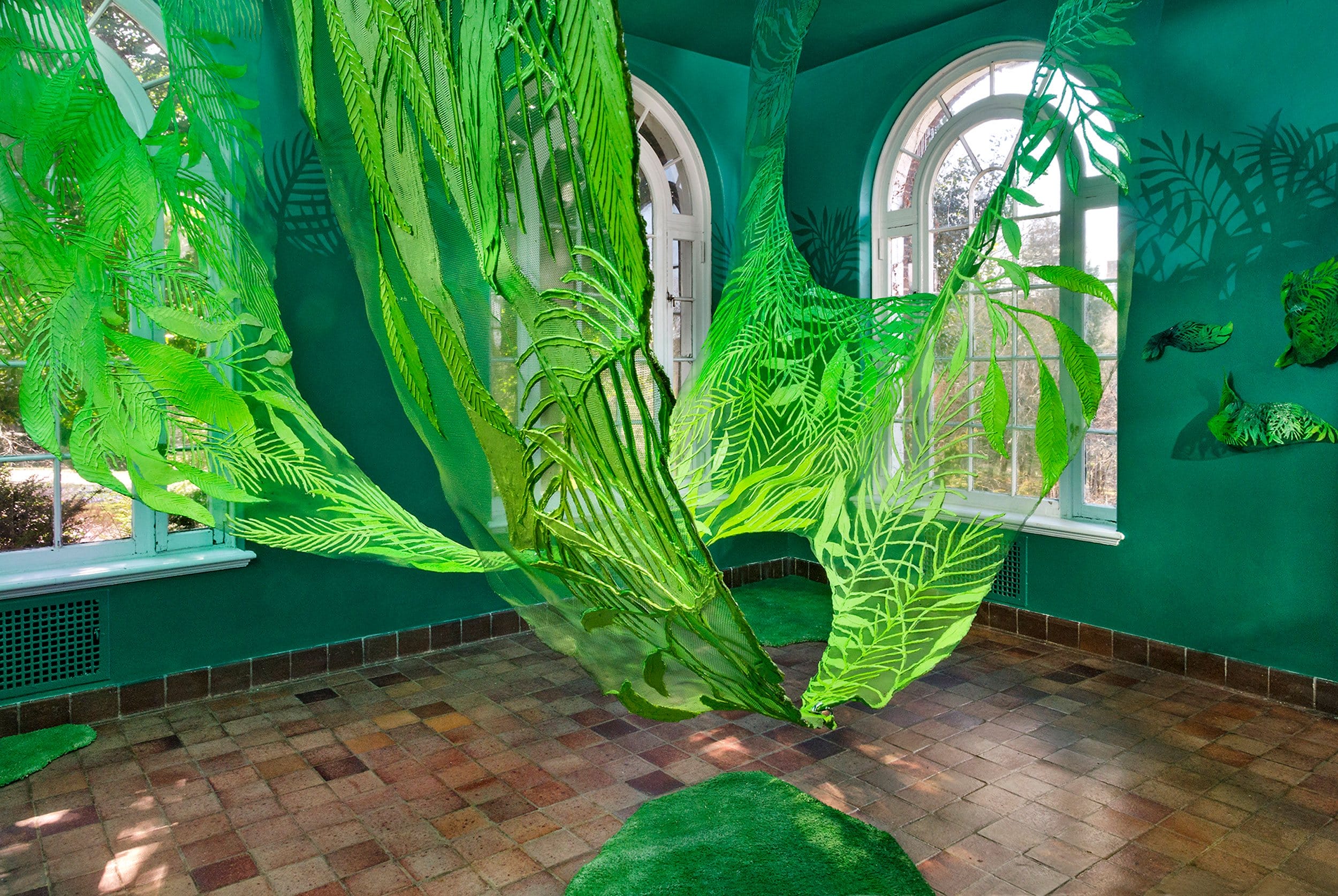 Room with green fabric art installation resembling leaves, featuring arched windows and brick flooring.