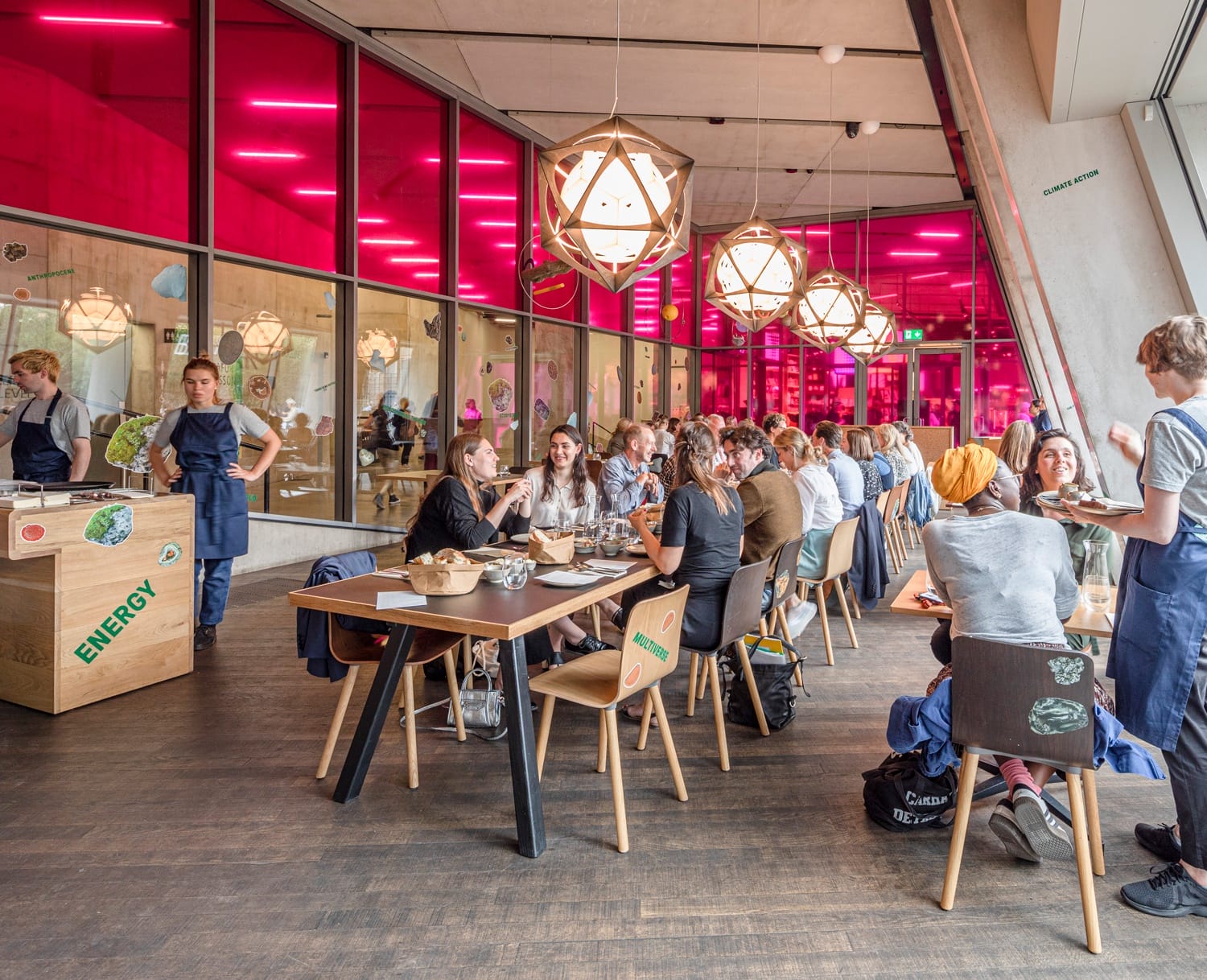 People dining in a modern restaurant with red-lit windows and geometric pendant lights.