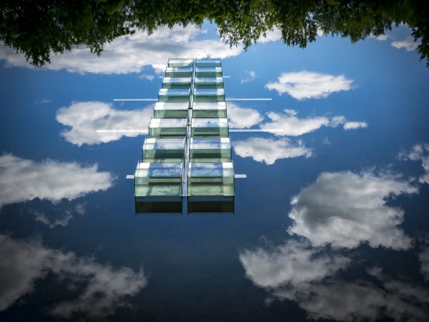 A modern glass structure reflecting against a calm body of water with clouds and trees in the background.