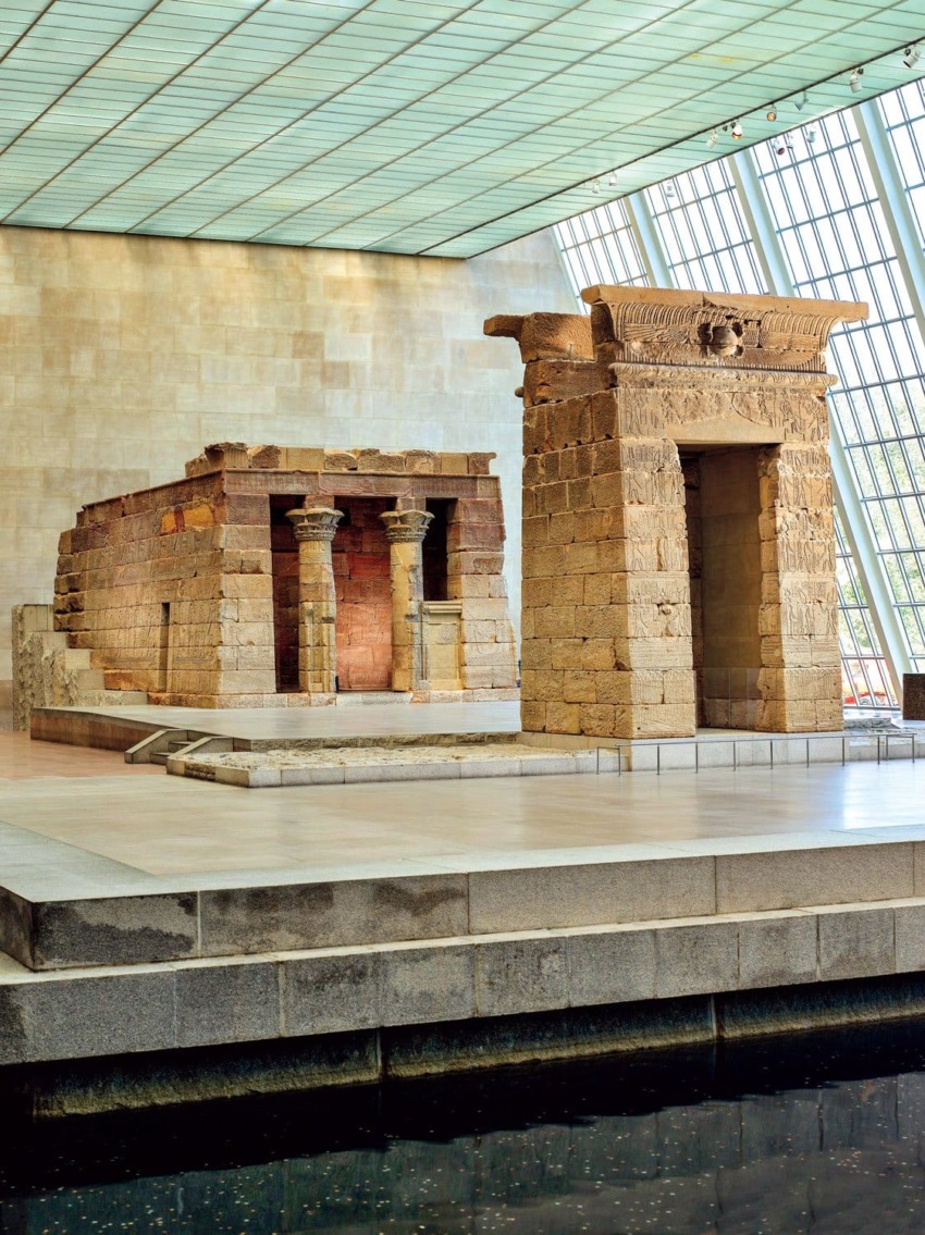 Ancient stone temple with columns inside a modern museum setting, surrounded by water, large windows in the background