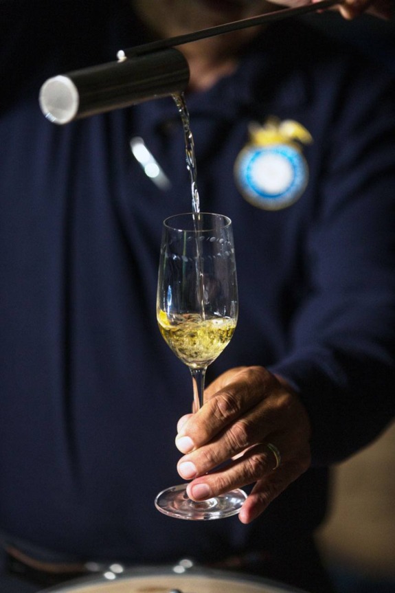 Person pouring clear liquid into a wine glass from a spout, wearing a navy blue shirt with a logo.