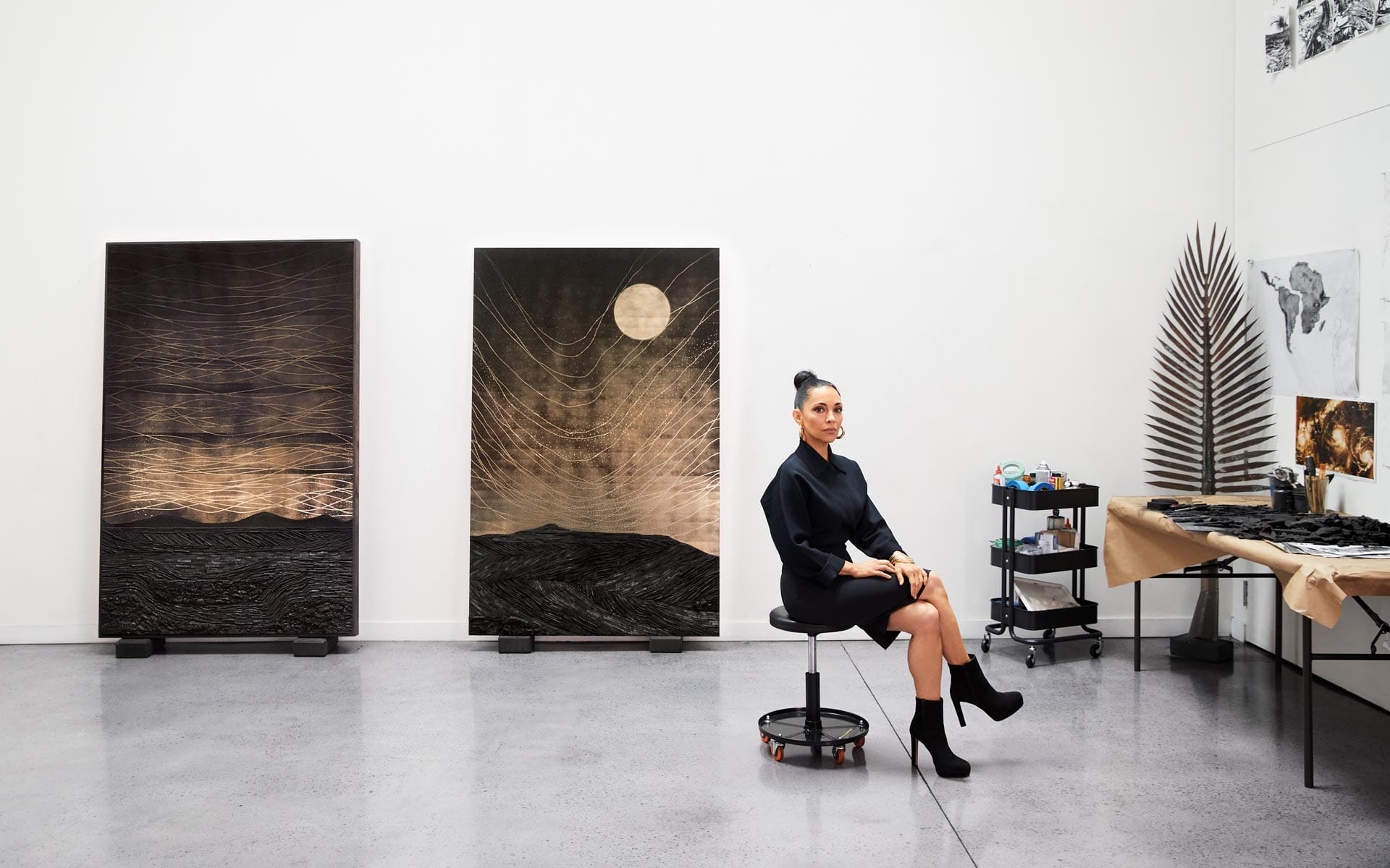 Artist seated in a studio with abstract paintings, wearing a black outfit, surrounded by art supplies on a white wall background.