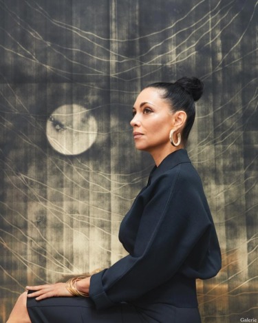 Person sitting sideways in front of an abstract moon and web-patterned backdrop, wearing elegant black attire and gold jewelry.