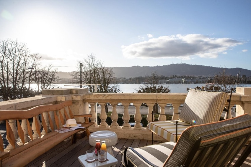 Sunny balcony with chairs, drinks, and a hat overlooks a scenic lake and mountain view under a partly cloudy sky.