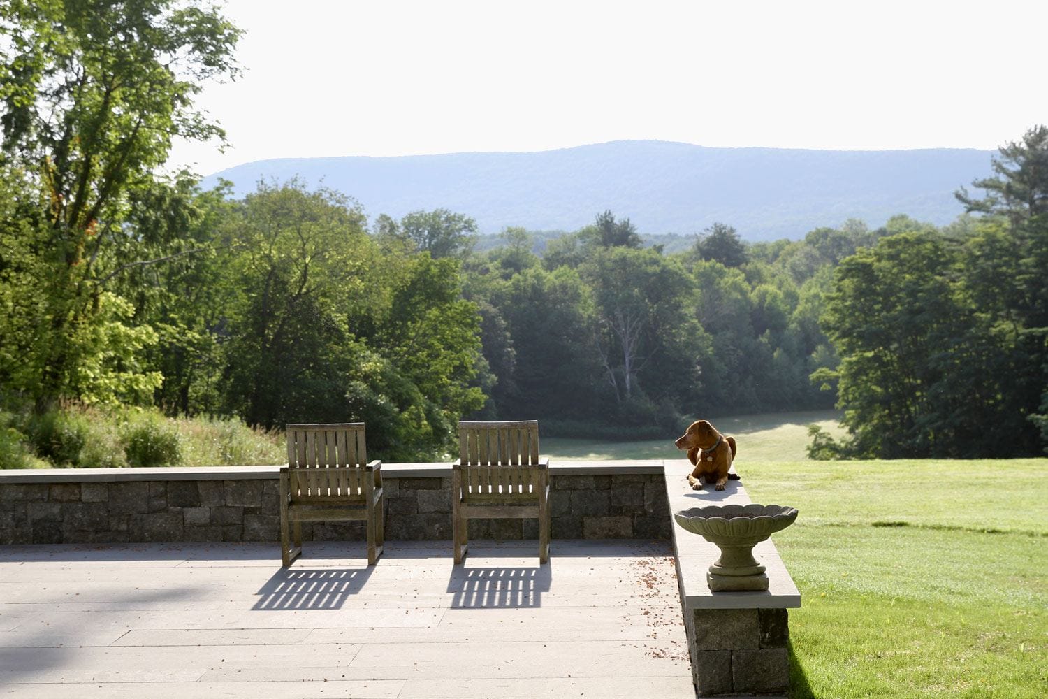 Two wooden chairs on a stone patio overlooking a green landscape with a toy dog sitting on a ledge.