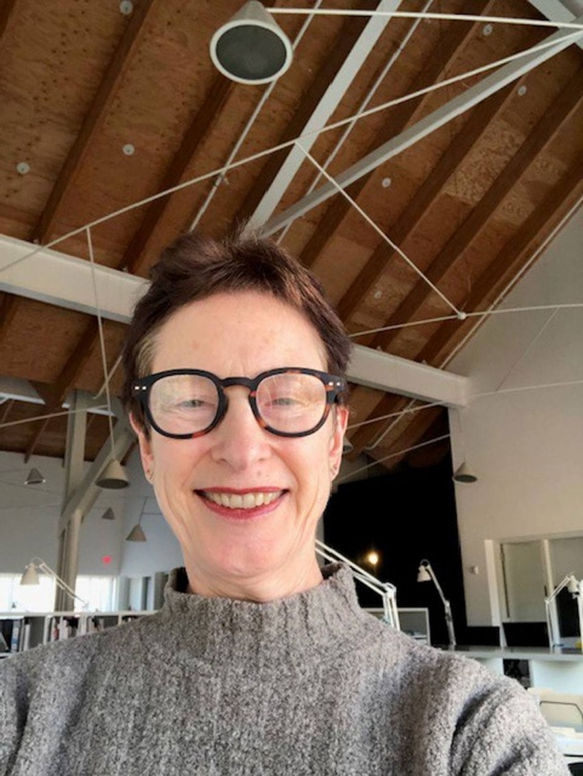 Person wearing glasses and a gray sweater smiling in a modern workspace with wooden ceilings and hanging lights.