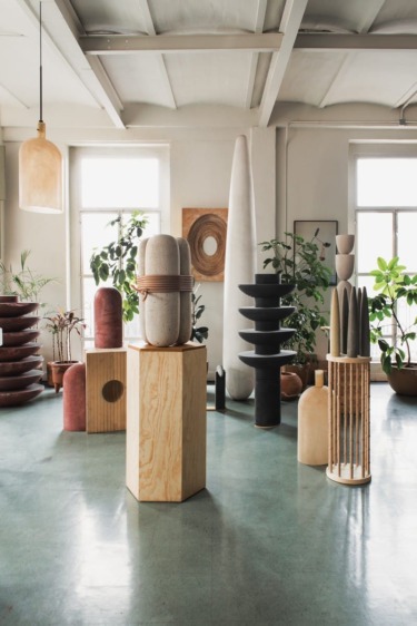 Room with modern sculptures and plants, featuring tall, abstract forms and natural light from large windows.