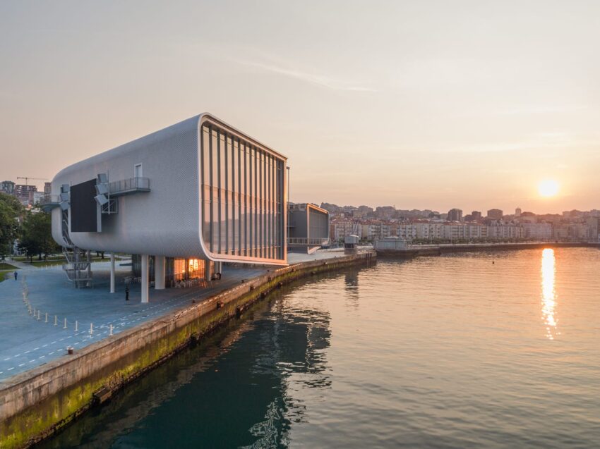 Modern waterfront building with large windows at sunset, overlooking calm water and distant cityscape.