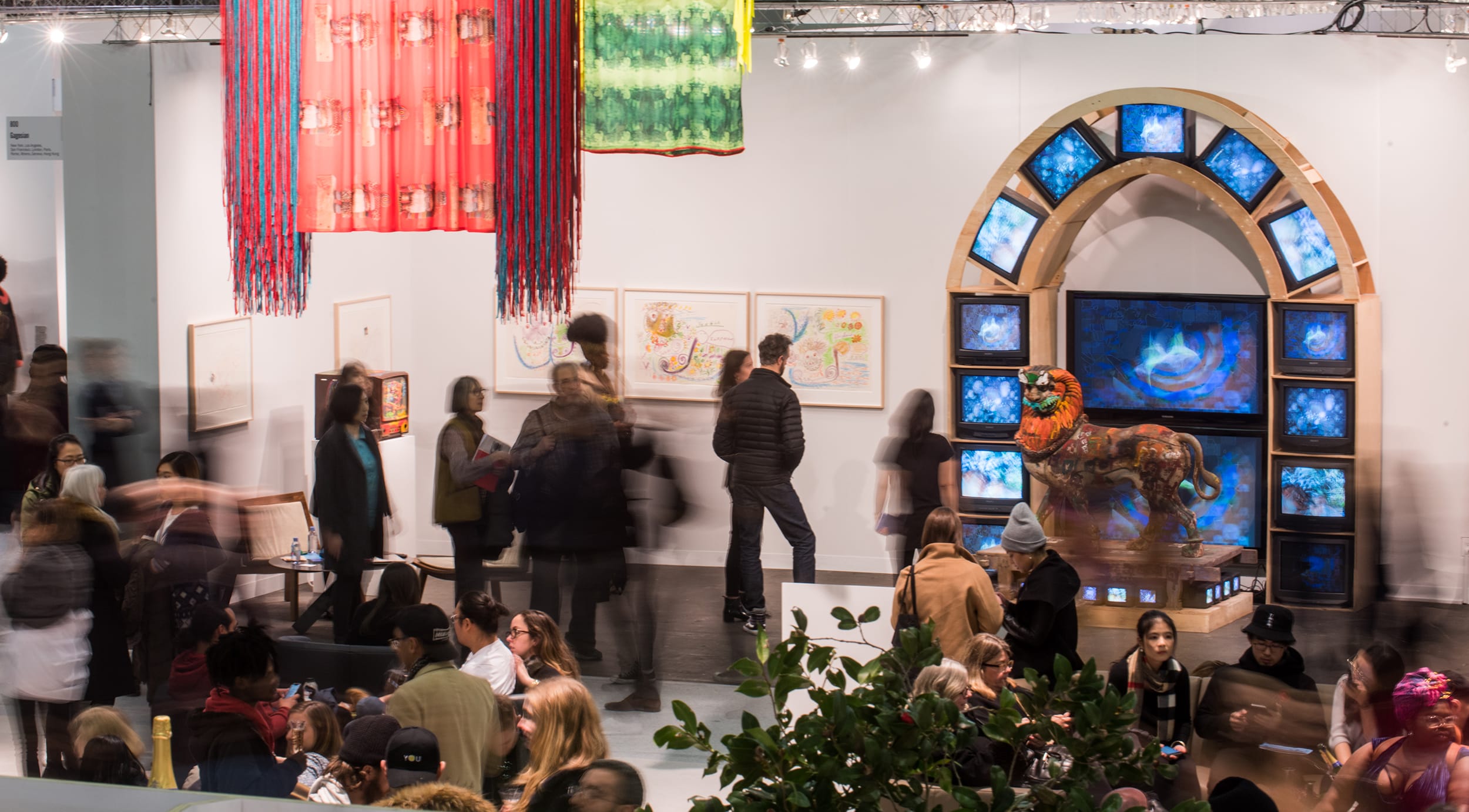 Art gallery with vibrant installations, including a digital lion sculpture and colorful hanging fabric, attended by numerous visitors.