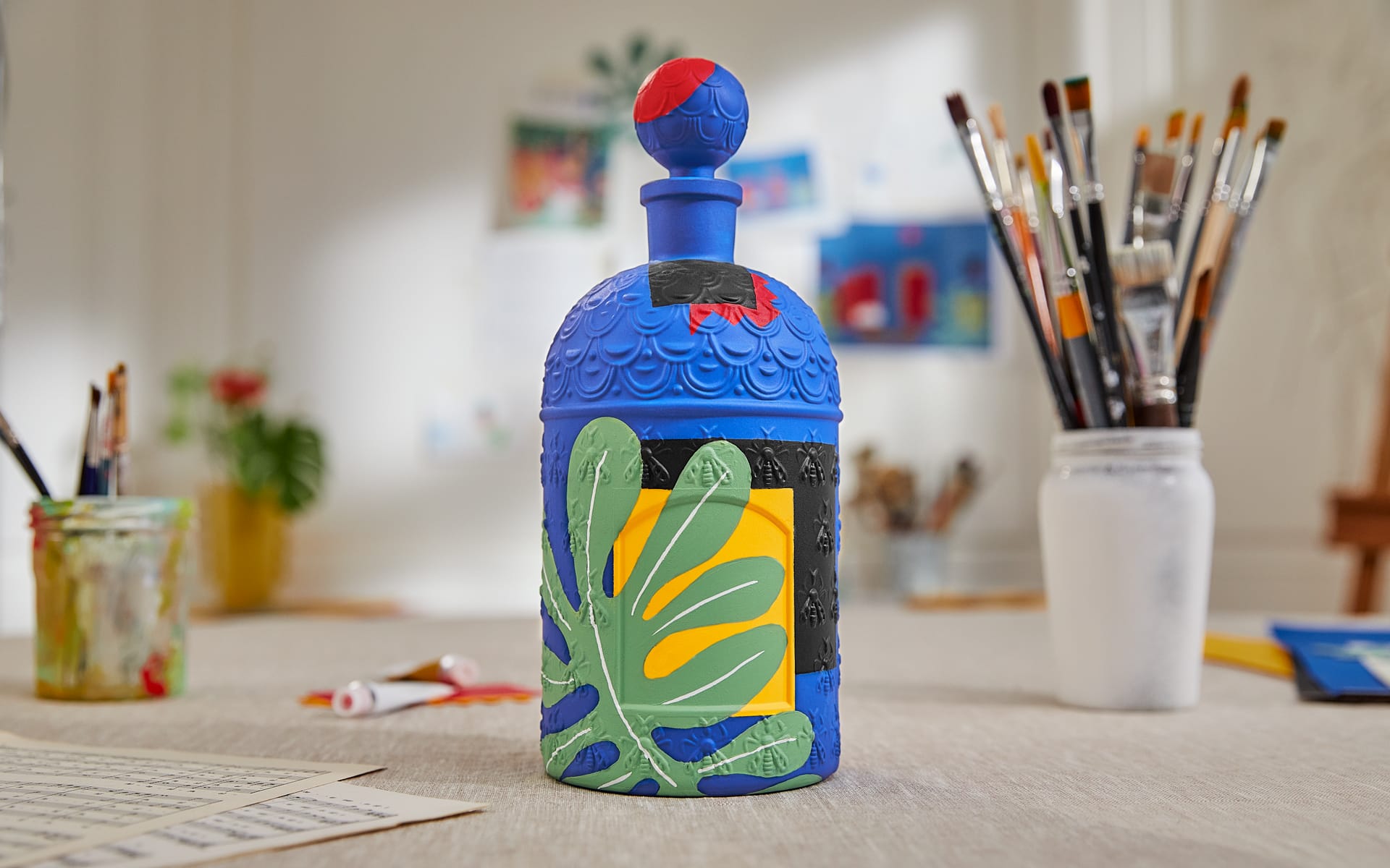 Colorful, intricately designed blue bottle with leaf patterns on a table surrounded by art supplies and brushes.