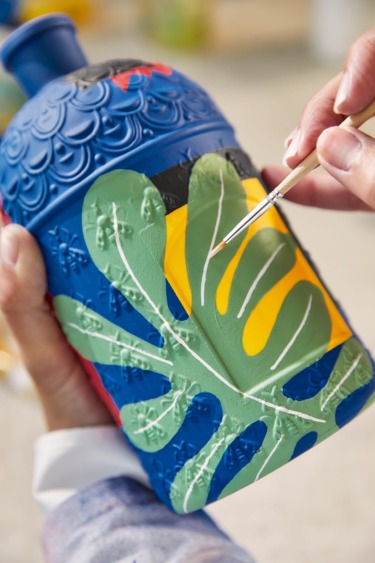 Person painting colorful design with leaves on a blue decorative bottle using a fine brush.