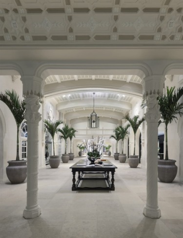 Elegant interior with ornate columns, large potted plants, and a central table under a high ceiling in a spacious hallway.