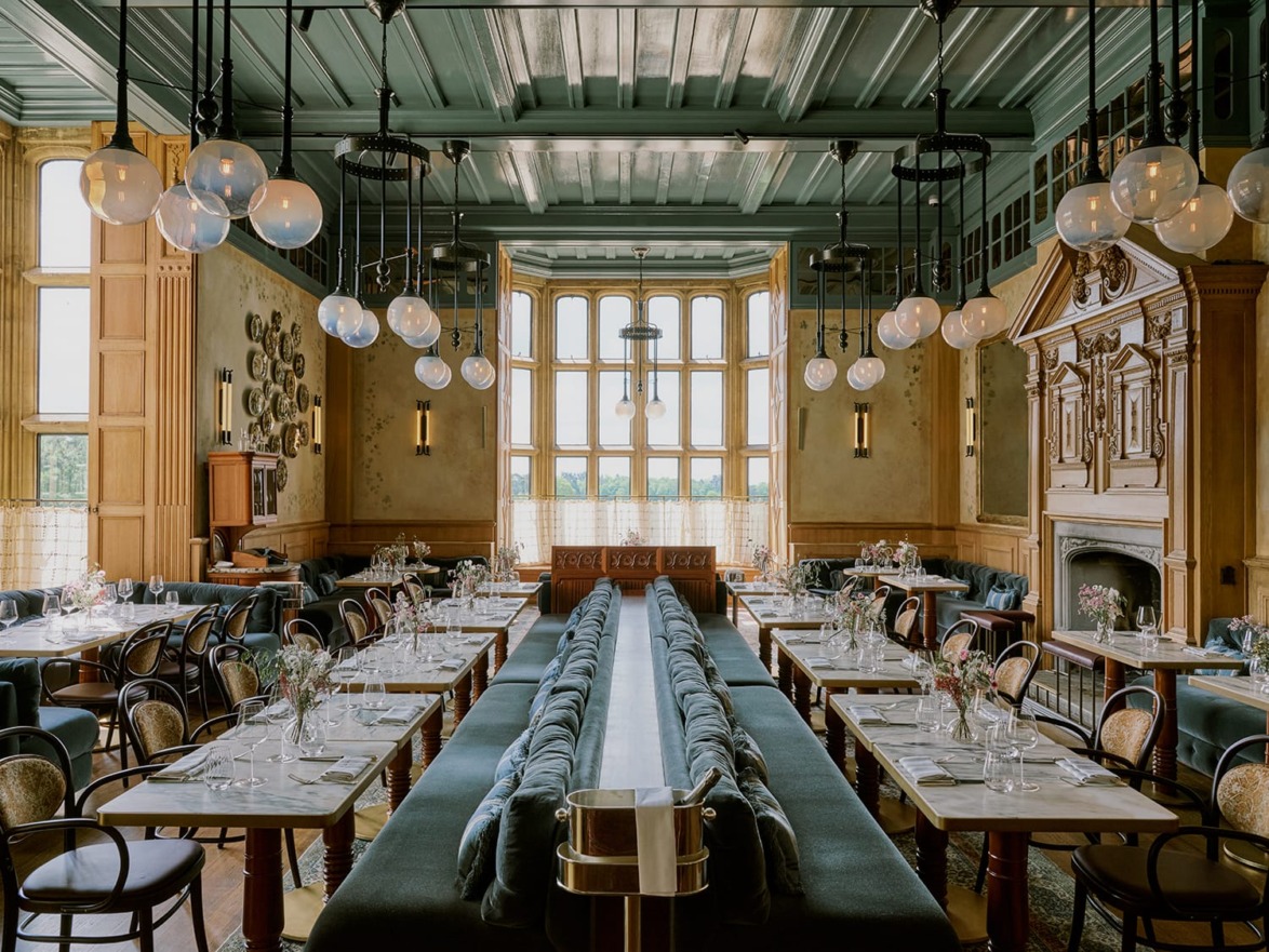 Elegant restaurant interior with long tables, cushioned seating, hanging globe lights, and large windows.