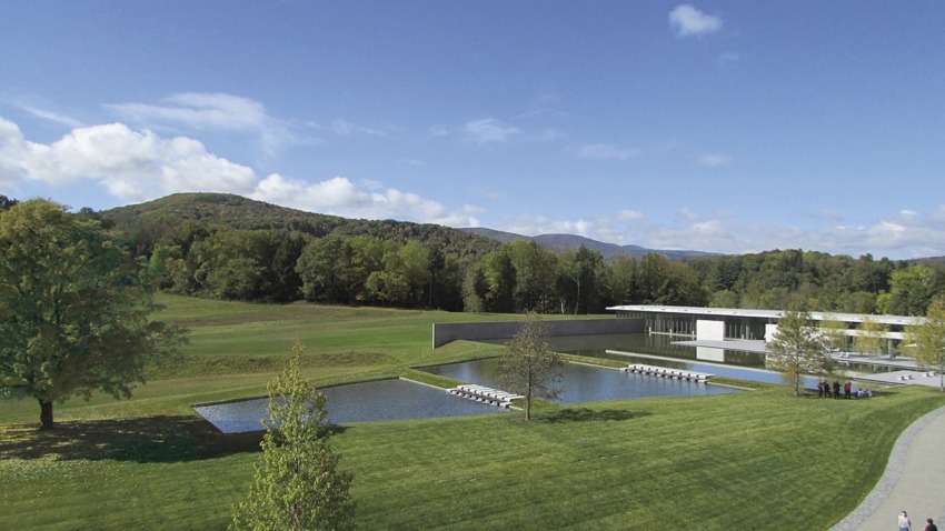 Modern art museum with a reflecting pool and lush green landscape, set against a backdrop of rolling hills and a blue sky.