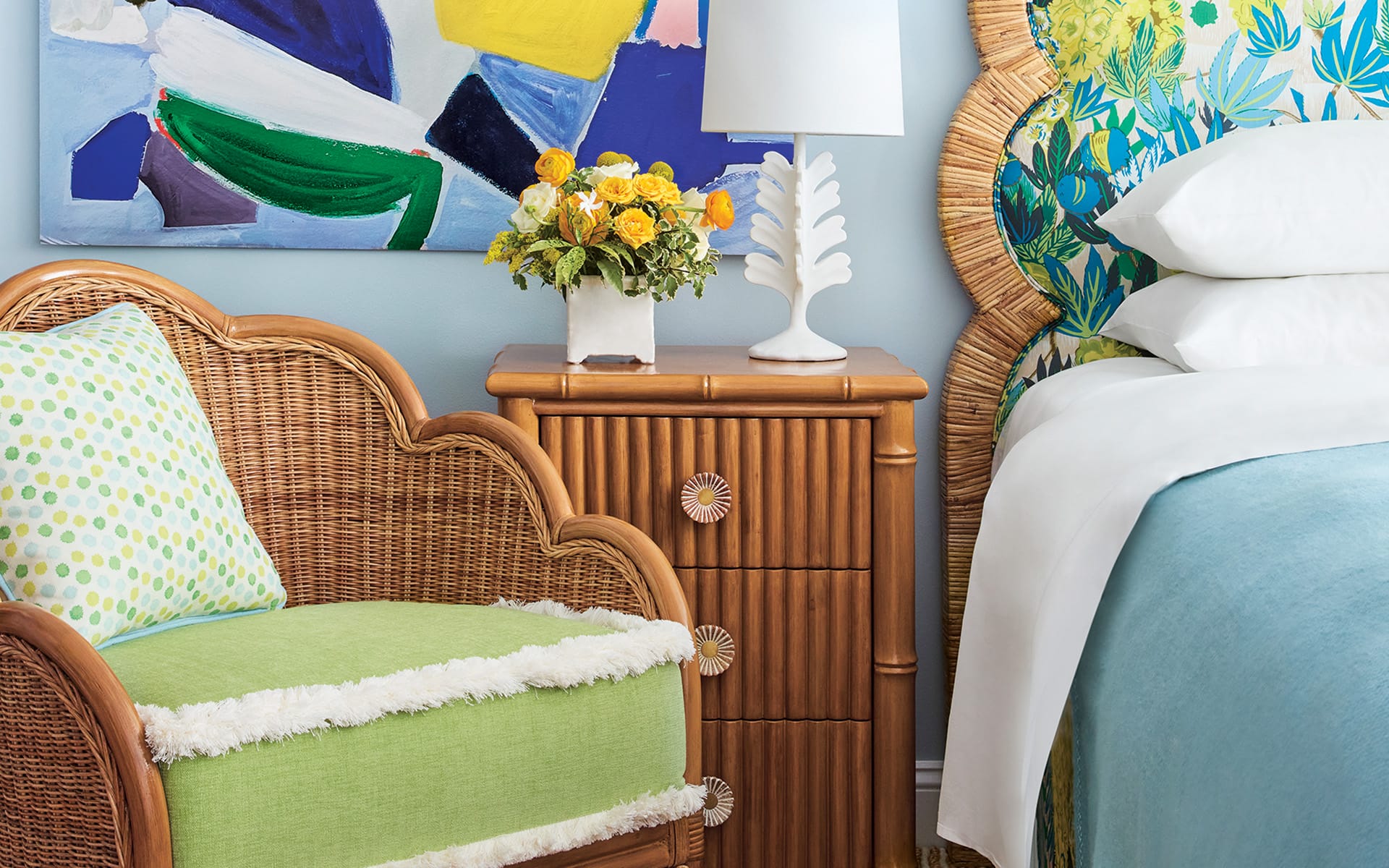 Tropical-themed bedroom with wicker chair, vibrant floral artwork, and colorful bed linens next to a wooden nightstand with flowers.