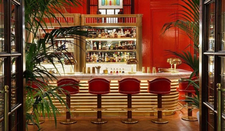 Art deco bar with red walls, gold accents, and four red stools, surrounded by lush green plants.