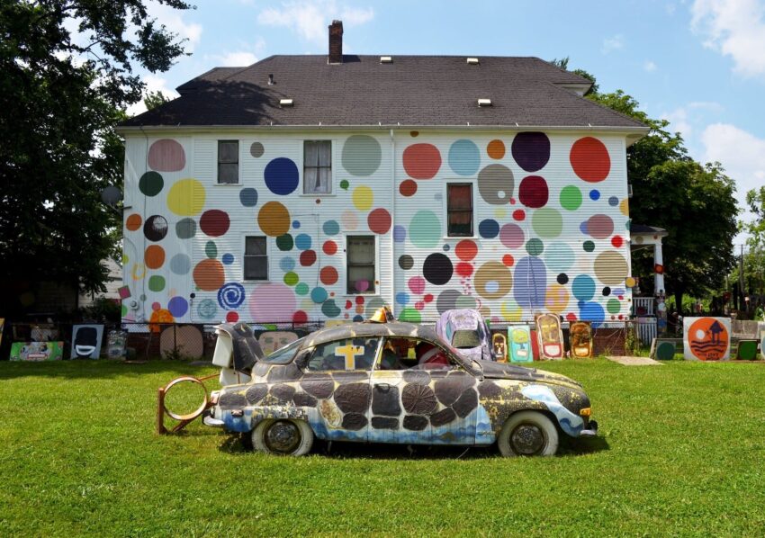 Colorful mural with circles on house exterior, artistic old car covered in patterns, vibrant grass, and trees in the background.