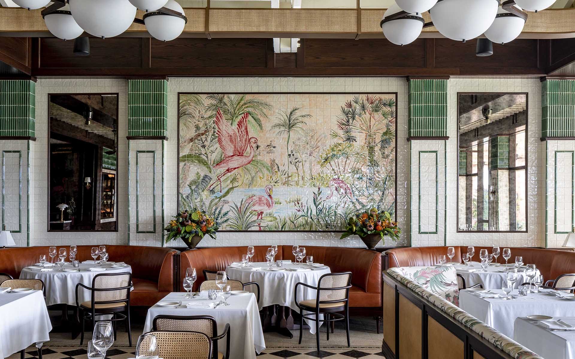 Elegant restaurant interior with white tablecloths, leather seating, a large tropical mural, and decorative lighting fixtures.