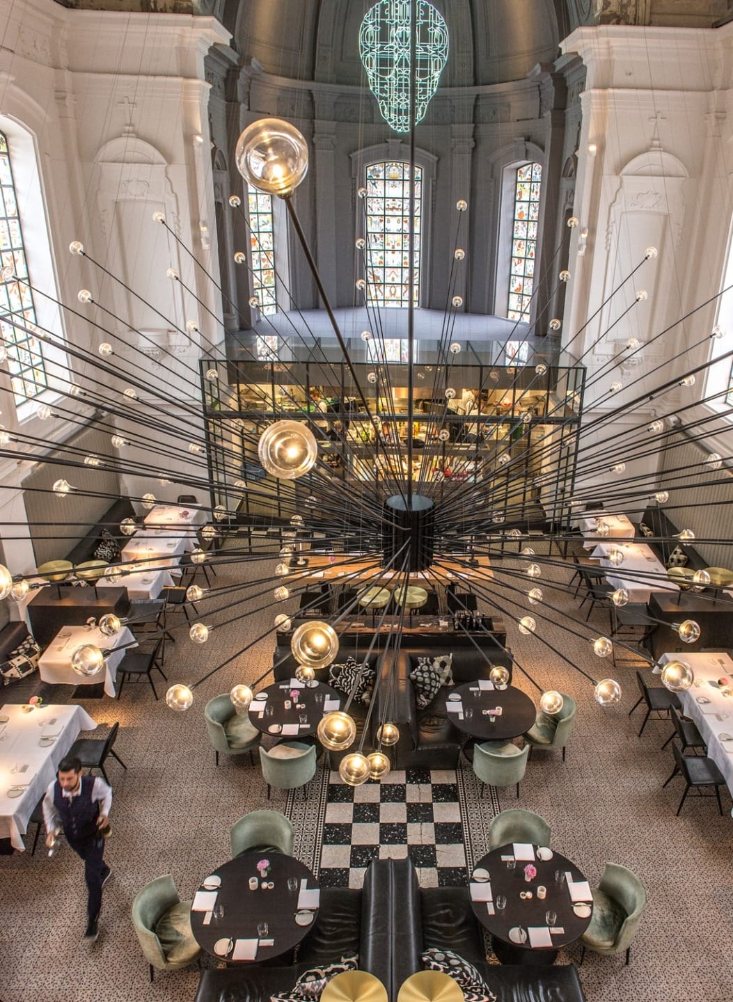 Modern restaurant interior with large chandelier, round tables, and a checkered floor, viewed from above.